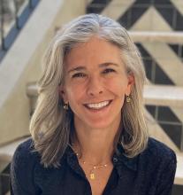 Tanya Clement, with shoulder-length gray hair and a black shirt, smiles while wearing gold accessories and sitting in front of a staircase.