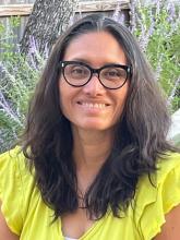 A woman wearing black glasses and a yellow blouse smiles while standing outside.