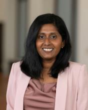 Portrait of a woman with straight black hair, wearing a light pink blazer and mauve top, smiling in a professional indoor setting.