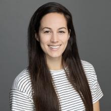 Professional portrait of a woman with long dark hair smiling at the camera against a gray background, wearing a white and black striped top