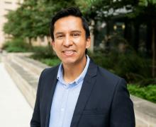 Professional headshot of a man with short black hair wearing a navy blazer over a light blue button-up shirt, smiling against a background of greenery and a walkway.