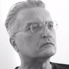 Black and white close-up portrait of an older person with swept-back hair wearing round wire-frame glasses, looking off to the side with a serious expression.