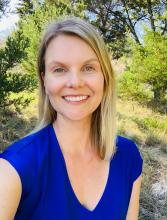  Smiling woman with straight light brown hair wearing a red floral dress, standing outdoors in front of trees and greenery.