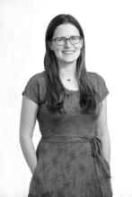 Black and white portrait of a smiling woman with long straight hair and glasses, wearing a patterned dress with hands in pockets.