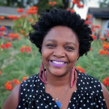 Smiling woman with short natural hair and purple lipstick, wearing polka-dot top and statement earrings, standing outdoors with vibrant orange flowers in the background