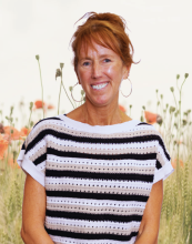 A woman with red hair in a loose updo stands smiling in front of a soft-focus background of wildflowers and wheat. She is wearing a short-sleeved, striped knit top and hoop earrings.