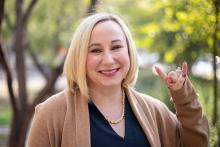 A smiling woman with chin-length blonde hair stands outdoors in front of blurred trees. She is wearing a camel-colored cardigan over a dark top, a gold chain necklace, and is making the ‘Hook ’em Horns’ hand gesture with her right hand.