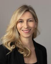 A professional portrait of a woman with long, wavy blonde hair wearing a black blazer. She is smiling warmly at the camera against a neutral gray background.
