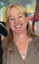 A woman with straight blonde hair smiles broadly in front of a colorful, abstract background. She is wearing a taupe blouse and two necklaces—one with a starfish-shaped pendant and another.