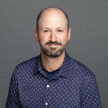 A headshot of Steve Finkelstein, a white man with a dark brown mustache and beard, in a blue button up dress shirt.