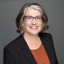A headshot of Kristin Patterson, a white woman with dark brown and blonde hair, smiling in a blazer and orange shirt.