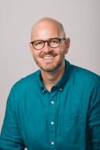 The head and torso of Matt Bowers, a white man with a brown mustache and beard, smiling in a teal button up shirt.