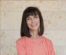 Headshot photo of a white woman with dark brown, shoulder-length hair standing in front of a beige stone wall. Her body is turned 45 degrees away from the viewer but looking directly at the camera and smiling.