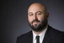 A headshot of Tolga Ozyurtcu, a man with a dark brown beard, smiling in a suit and tie.