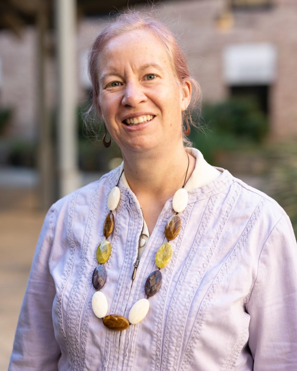 A woman wearing blouse and a beaded necklace, is smiling while standing outdoors.