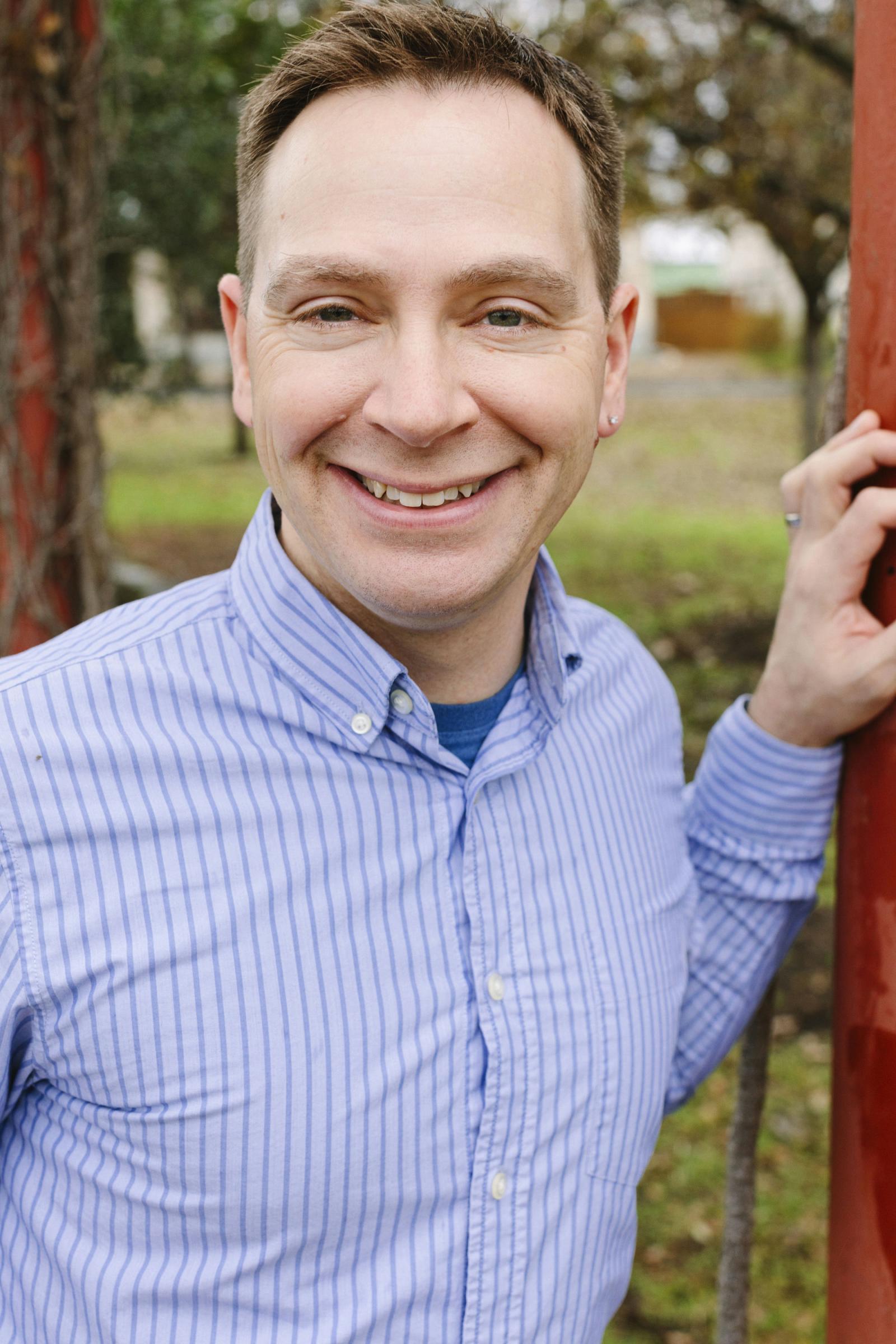 Elon Lang, wearing a vertically blue-striped shirt, smiles while leaning against a red pole.
