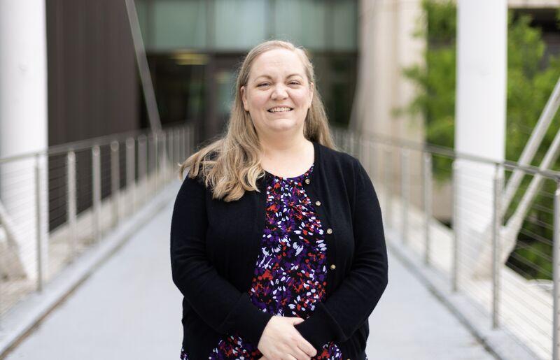 Smiling woman with medium blonde hair wearing a black cardigan and floral top, standing on a CMA bridge.