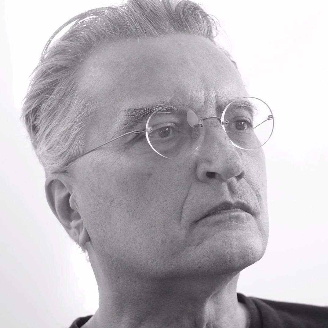 Black and white close-up portrait of an older person with swept-back hair wearing round wire-frame glasses, looking off to the side with a serious expression.