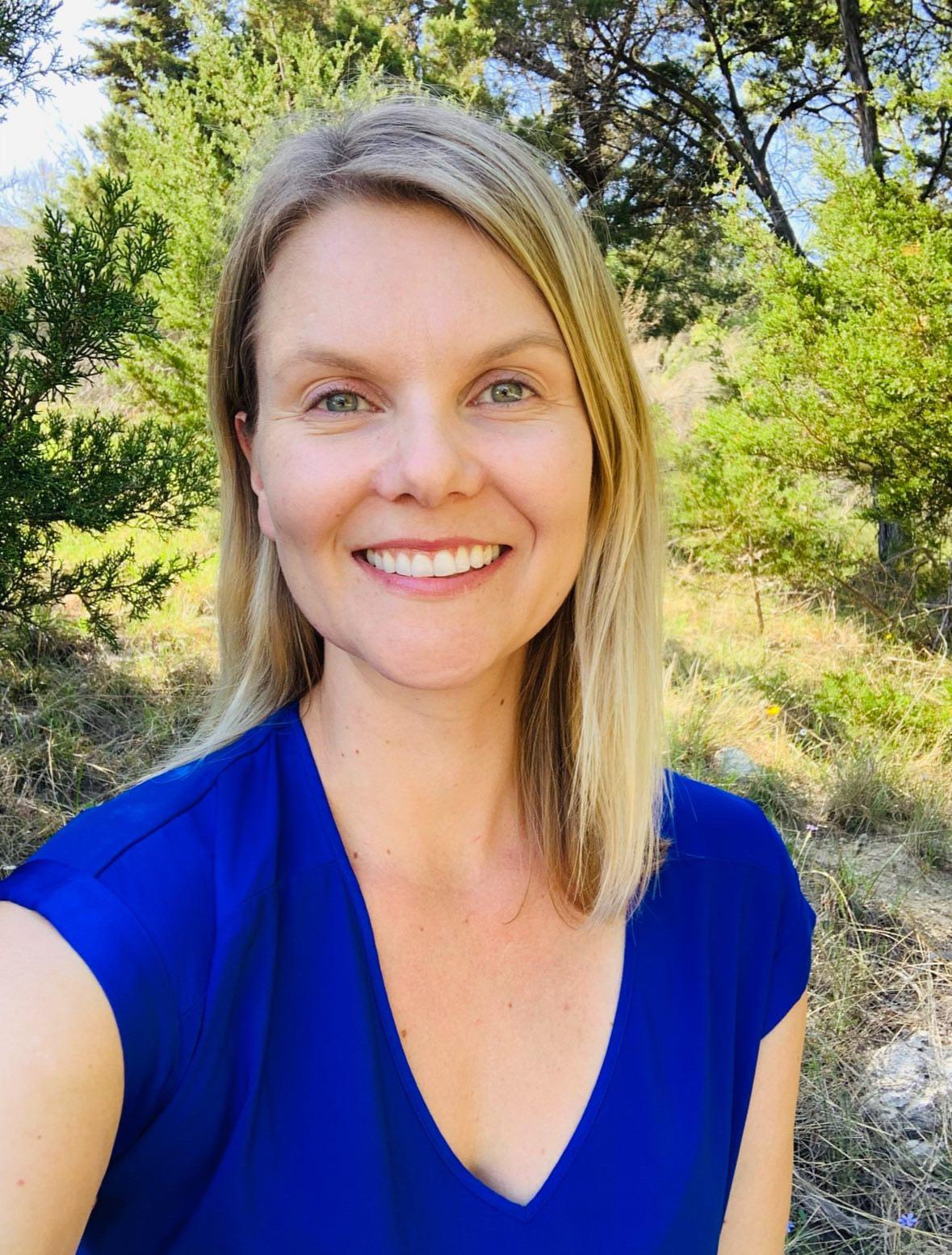  Smiling woman with straight light brown hair wearing a red floral dress, standing outdoors in front of trees and greenery.