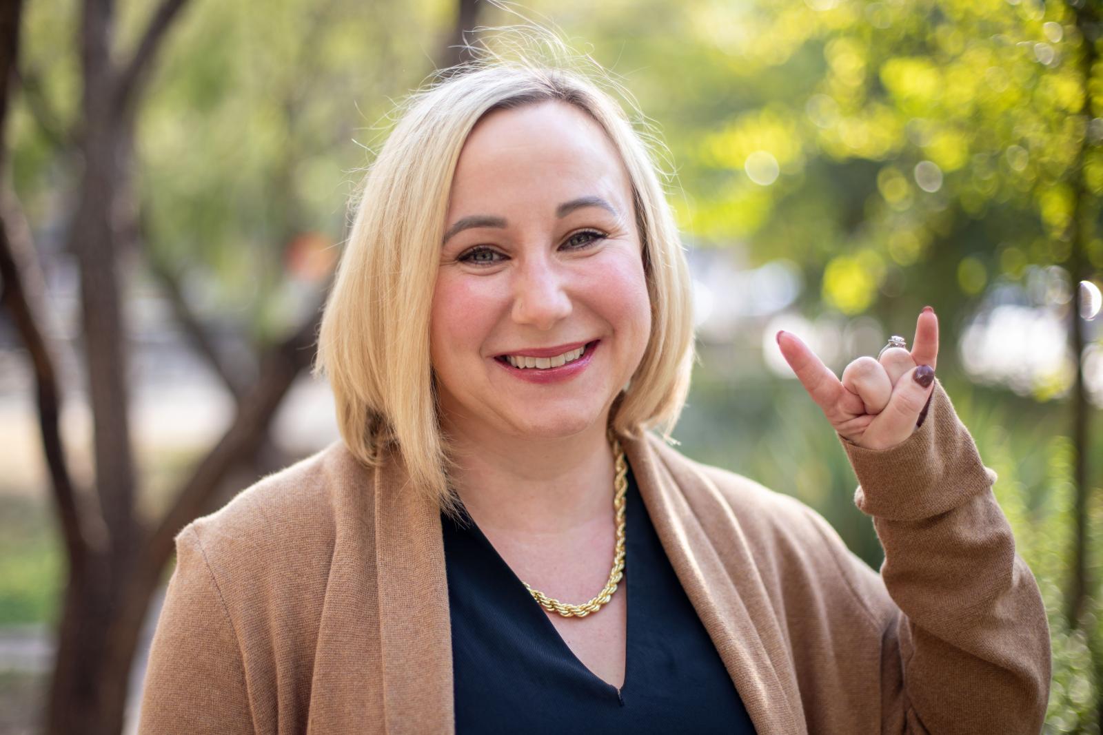 A smiling woman with chin-length blonde hair stands outdoors in front of blurred trees. She is wearing a camel-colored cardigan over a dark top, a gold chain necklace, and is making the ‘Hook ’em Horns’ hand gesture with her right hand.