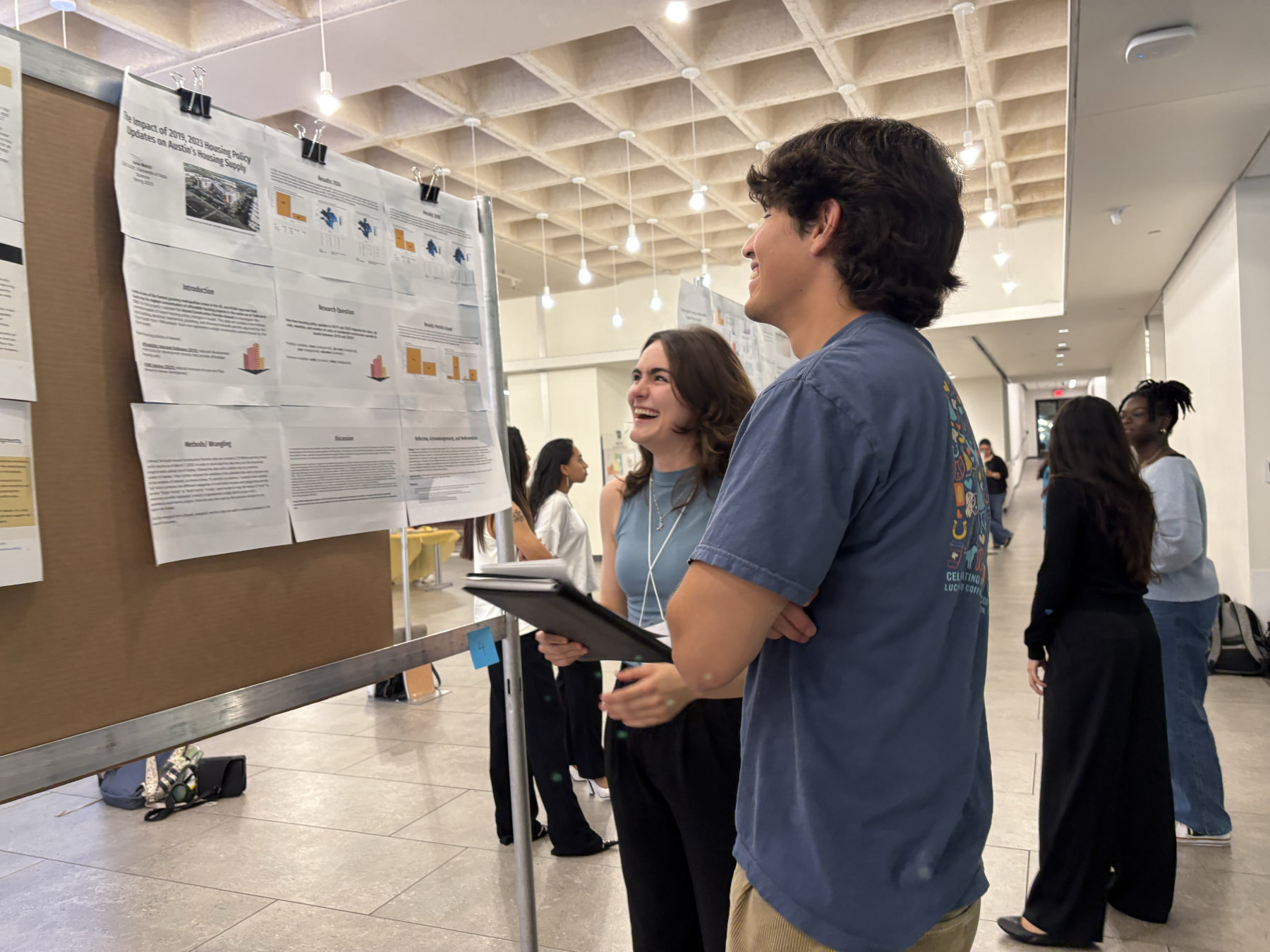Two people stand in front of a research poster, smiling and talking during a presentation session in a bright hallway.