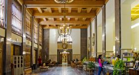 Large rectangular room with high ceilings and chandeliers and various individuals sitting and standing in the distance