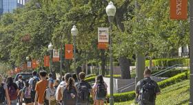 Students traveling along a pediastrian walkway with lamp posts and UT branded flags. Green grass and green grass appear above the and to the right of the students
